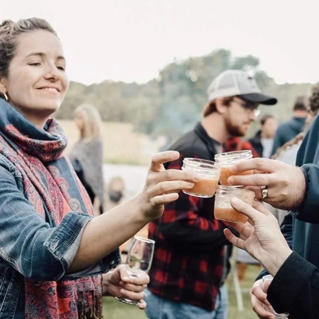 A woman in a scarf and denim jacket at The Roost Farm's outdoor Farm to Table event clinks glasses (in this case, a jar of maple syrup based drink) with someone outside the frame of the image. Several people are mingling in the background.