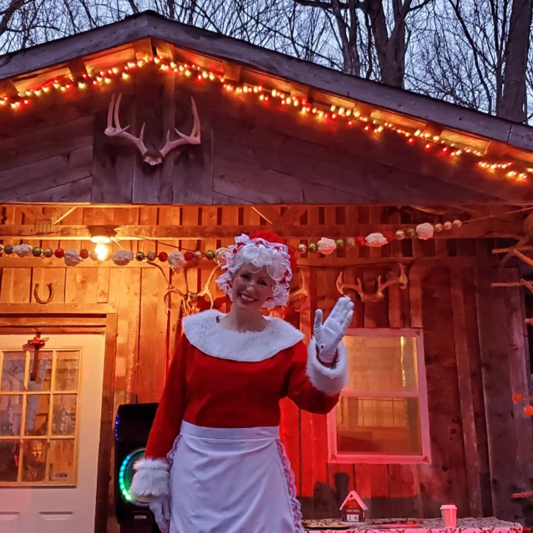 A smiling Mrs. Claus waves in front of a maple sugar shack decorated for the holiday season, at The Roost's Yulefest on the Farm event.